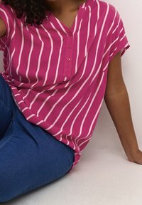 Blouse rose à manches courtes avec des rayures blanches verticales, dotée d'un col en V peu profond et d'un détail à boutons. Associée à un jean bleu.