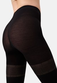 Primo piano di una persona che indossa collant neri a fantasia, evidenziando la texture e il design intorno alla coscia e ai fianchi.