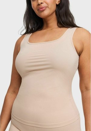 Vrouw met een huidtint van gemiddelde toon die een mouwloze beige tanktop draagt, zichtbaar van halverwege de borst tot de heupen tegen een effen achtergrond.