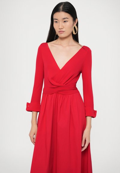 Piros átöltős ruha mély V-nyakkivágással, hosszú ujjakkal és testhezálló derékkal. Díszítik omló redők és begombolható ujjszegélyek. Simább szövetű anyag.