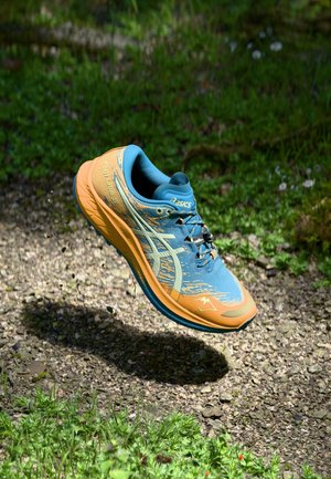 Zapatilla de correr Asics azul y naranja flotando sobre un sendero de grava con hierba verde y plantas de fondo.