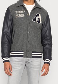 Grå ullblandad varsityjacka med läderärmar, broderad bröstlogotyp, randiga ribbade muddärmar och nederkant, tryckknappsstängning, två sidofickor.