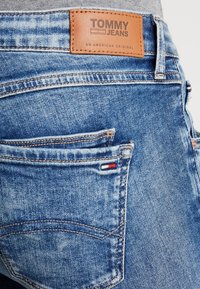 Jeans de mezclilla en azul claro, con una etiqueta de cuero marrón de la marca que dice "TOMMY JEANS" y un pequeño logo rojo, blanco y azul en el bolsillo.