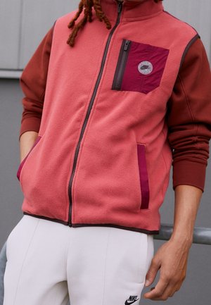 Osoba ubrana w czerwoną i różową bluzę polarową z suwakiem, z bordową kieszenią na piersi i białe spodnie Nike, stojąca z jedną ręką w kieszeni.