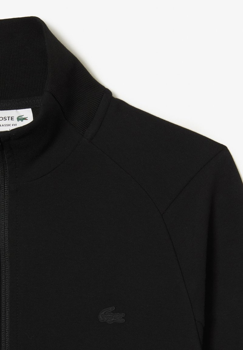 Giacca nera con zip e colletto alto, realizzata in tessuto strutturato. Presenta un logo in rilievo discreto sul petto e dettagli delle cuciture a contrasto.