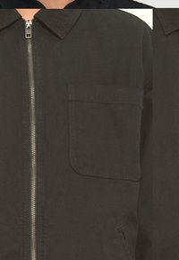 Giacca in cotone nera con zip frontale, colletto a punta e taschino sul petto. Il tessuto ha una finitura opaca e dettagli di cucitura minimi.