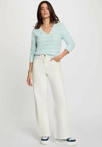 Maglione azzurro chiaro a collo a V con motivo a zigzag, abbinato a jeans bianchi a vita alta e gamba larga e sneaker con dettagli blu.