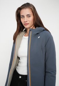 Gray insulated jacket with a hood, contrasting beige lining, and a multicolored zipper detail. Features a small logo on the chest.