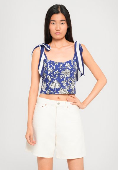 Woman wearing a blue floral crop top with striped shoulder ties and high-waisted white shorts, standing with one hand on hip.