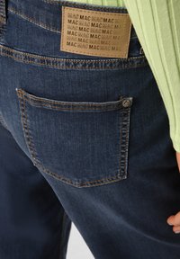 Dark blue denim jeans with a textured surface, featuring a square back pocket and a tan label stitched near the waistband.