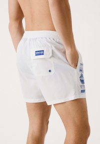 Bañador blanco con cintura elástica, con un bolsillo trasero y un logo de Red Bull Racing en azul. Textura suave y diseño minimalista.