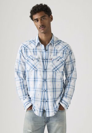 Mann trägt weißes und blaues kariertes Hemd mit Brusttaschen, steht mit den Händen in den Taschen vor einem einfarbigen Hintergrund.