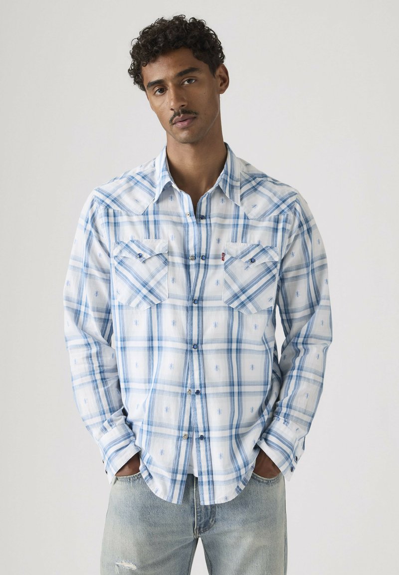 Mand iført hvid og blå plaid skjorte med brystlommer, stående med hænderne i lommerne mod en ensfarvet baggrund.