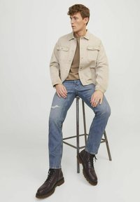 Chaqueta de mezclilla beige sobre una blusa marrón, combinada con jeans claros rasgados y botas de cuero marrón oscuro con cordones. Los taburetes añaden un contraste en la altura.