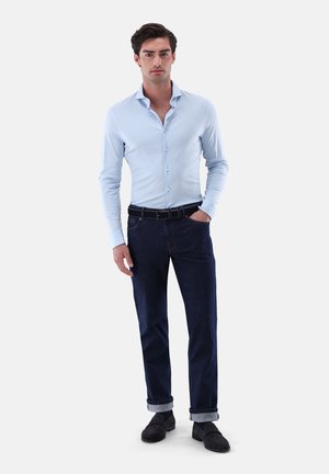 Hellblaues langärmeliges Hemd mit Knöpfen, dunkelblaue Jeans mit Umschlägen, schwarzer Gürtel und schwarze Schuhe, vor einem weißen Hintergrund.
