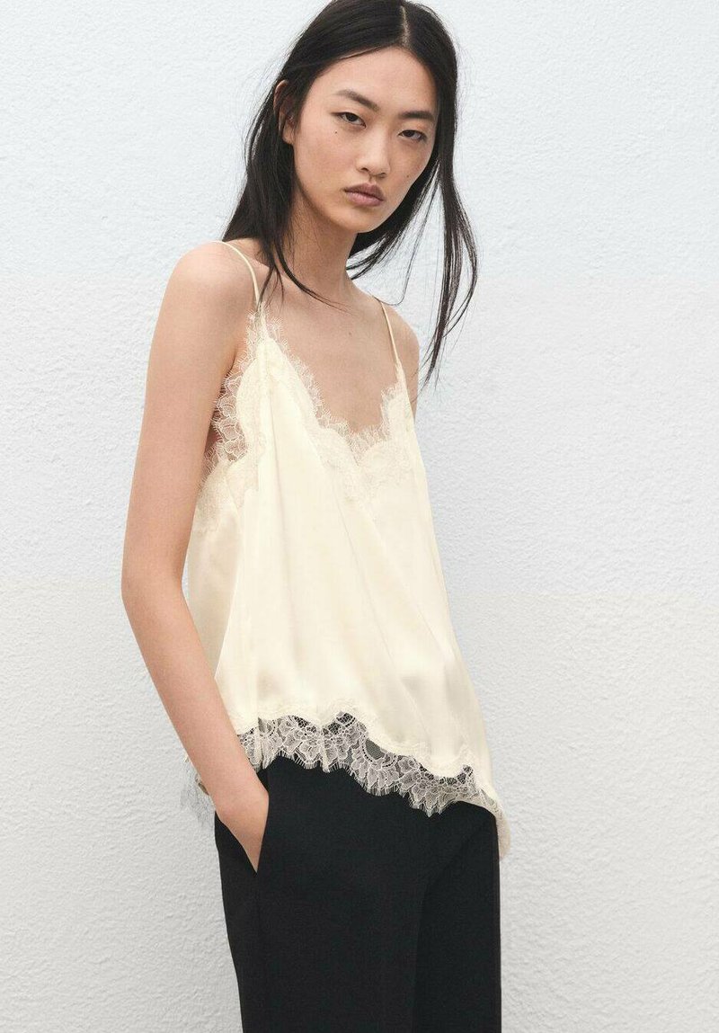 Camisola de seda en amarillo pálido con ribete de encaje en el escote y el dobladillo. Presenta tirantes finos y un ajuste relajado. Combinada con pantalones negros.