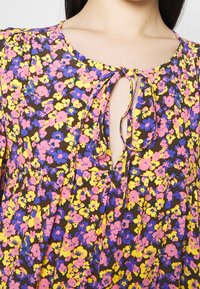 Blommig klänning med brun bakgrund som har rosa, gula och blå blommor. Nyckelhålsnacke med knytDetalj och veckad kjoldesign.