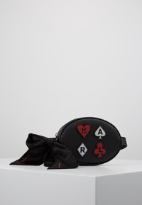 Svart oval läderväska med lappar av spelkortssymboler och bokstäver, med en stor svart rosett knuten på sidan, på en vit yta.