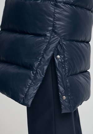 Cappotto imbottito blu navy con texture trapuntata, dotato di chiusure a scatto e una leggera apertura nell'orlo per migliorare il movimento e il design.