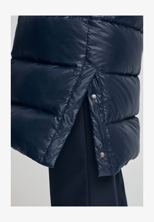 Abrigo de plumón azul marino con textura acolchada, que presenta cierres a presión y una pequeña abertura en el dobladillo para mejorar el movimiento y el diseño.