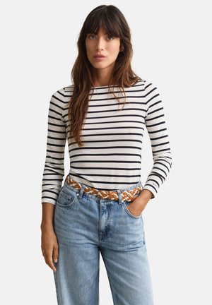 Femme portant un t-shirt à manches longues rayé blanc et noir, rentré dans un jean bleu avec une ceinture tressée marron, debout avec une main dans la poche.