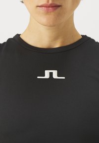 Juodas sportinis marškinėliai su lygia tekstūra ir baltu logotipu krūtinės centre. Apvalus iškirpimas, prisitaikantis dizainas.