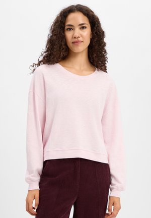 Junge Frau mit lockigem Haar, die ein hellrosa Langarmshirt und dunkelburgunderfarbene Cordhosen trägt, steht vor einem schlichten Hintergrund.
