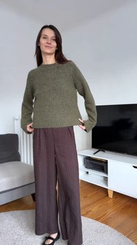 Olivovo zelený pletený sveter so voľným strihom, kombinovaný s voľnými širokými nohavicami tmavohnedej farby. Čierne tienisté topánky na drevenej podlahe.