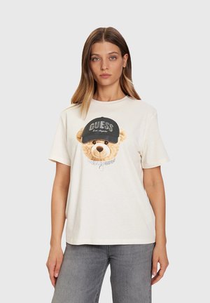 Donna che indossa una t-shirt bianca con la grafica di un orsetto che porta un berretto nero con la scritta "GUESS Los Angeles" e jeans grigi, in piedi davanti a uno sfondo semplice.