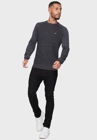 Dunkelgrauer Sweatshirt mit strukturierter Oberfläche und kontrastierendem Ärmel-Design, mit einem kleinen goldenen Logo auf der Brust, kombiniert mit schwarzen Jeans und weißen Turnschuhen.