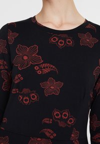 Robe noire à manches longues avec des motifs floraux rouges. Encolure ronde, tissu doux avec une texture lisse. Des motifs floraux clés ornent la surface.