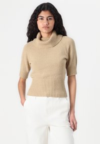 Beżowy sweter z krótkim rękawem z prążkowanym golfem i w talii. Materiał wydaje się miękki z teksturowanym wykończeniem, w połączeniu z białymi spodniami.
