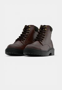 Braune Lederstiefel bis zur Knöchel mit schwarzen Gummisohlen, goldenen Ösen und Schnürdesign. Mit gepolstertem Kragen und strukturiertem schwarzen Außensohle.