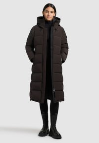 Frau in einem langen schwarzen gepolsterten Wintermantel mit Kapuze, schwarzen Leggings und klobigen schwarzen Stiefeln, die vor einem hellgrauen Hintergrund steht.