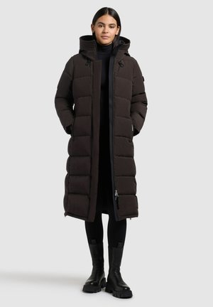 Vrouw in een lange zwarte gewatteerde winterjas met capuchon, zwarte leggings en stevige zwarte laarzen, staand tegen een lichtgrijze achtergrond.