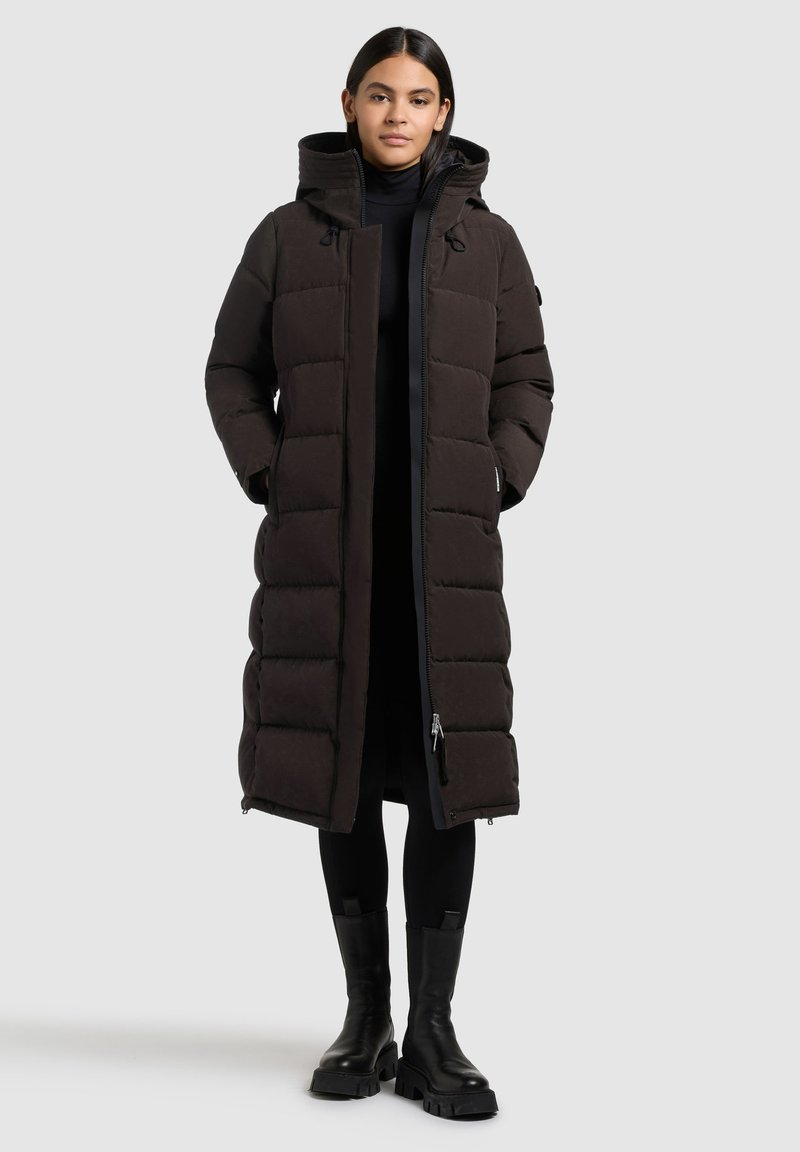 Frau in einem langen schwarzen gepolsterten Wintermantel mit Kapuze, schwarzen Leggings und klobigen schwarzen Stiefeln, die vor einem hellgrauen Hintergrund steht.
