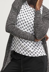 Un cardigan largo gris sobre una blusa blanca con un patrón de hojas negras, combinado con pantalones negros. Textura suave y ajuste relajado.