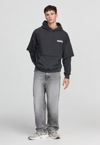 Felpa oversize grigio scuro con tasca frontale e logo, abbinata a jeans sfilacciati grigio chiaro e sneakers grigie con dettagli bianchi.