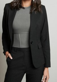 Schwarzer Blazer mit einem einzigen Knopf, kombiniert mit einem grauen Rippoberteil. Der Blazer zeichnet sich durch einen glatten Stoff, eine taillierte Passform und schmale Revers aus.
