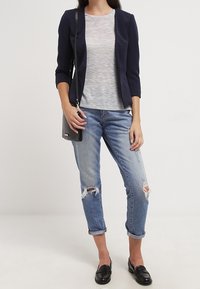 Marineblaue taillierte Blazer, hellgraues strukturierte Oberteil, ausgewaschene hellblaue Jeans, schwarze Loafers und eine kleine schwarze Umhängetasche.