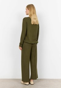Olivgrünes Oberteil mit langen Ärmeln in lockerer Passform, kombiniert mit weit geschnittenen Hosen, beide aus einem weichen Textilmaterial. Neutrale Schuhe vervollständigen das Outfit.