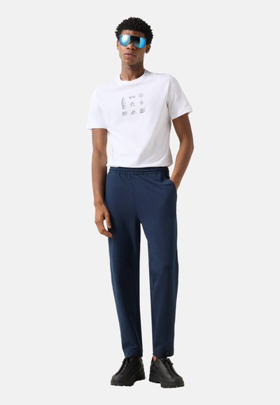 Jeune homme portant des lunettes de soleil bleues réfléchissantes, un t-shirt blanc à motif graphique, un pantalon bleu marine et des baskets noires, debout avec une main dans la poche.