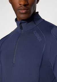 Marineblå genser med høy krage og kvart glidelås, med teksturert finish og sømdetaljer. "Bogner"-logo trykt på siden.