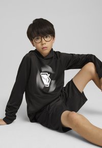 Sudadera deportiva negra con un diseño gráfico, combinada con pantalones cortos negros. Textura suave, forma ajustada y brillo sutil. Posado sentado.