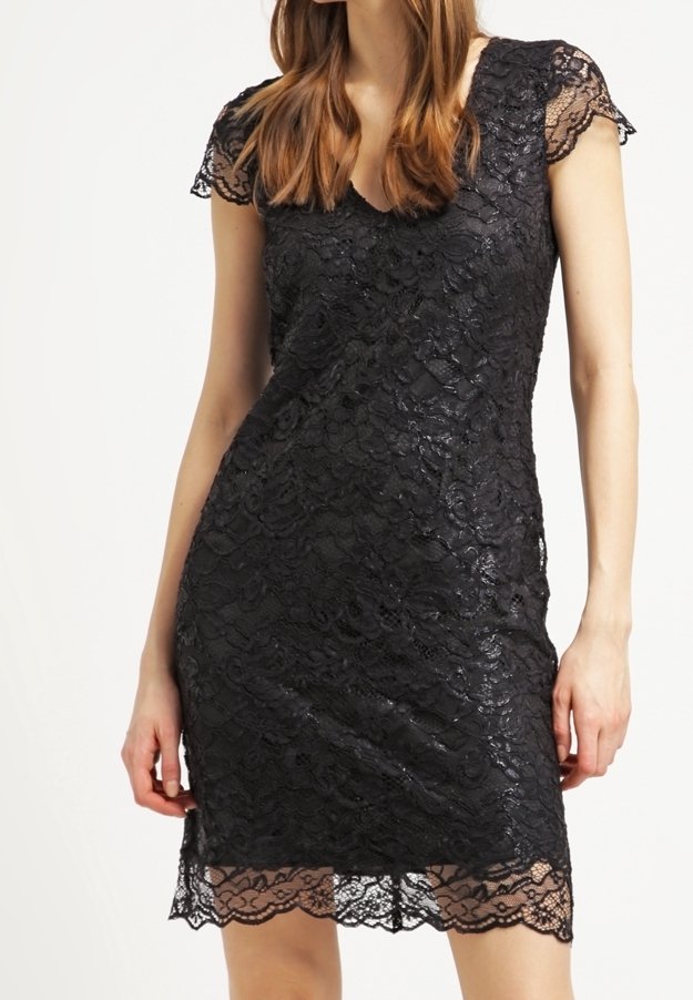 Kvinna iklädd en knälång svart spetsklänning med korta ärmar och vågig fåll, stående mot en vit bakgrund.