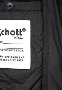 Hvidt stofmærke med sort tekst "Schott N.Y.C." syet ind i en sort quiltet jakke med hexagonal mønster og synlig lynlås.