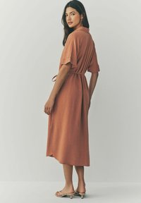 Robe midi en terracotta à manches courtes, taille ceinturée et détails plissés, fabriquée en tissu léger. Fente sur le côté et ourlet arrondi.