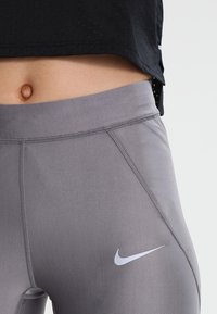 Szare legginsy sportowe o gładkiej fakturze, z kontrastowym białym logo Nike i szczegółowym przeszyciem na bocznych szwach.