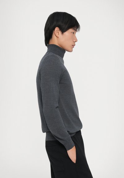 Junger Mann in grauem Rollkragenpullover und schwarzen Hosen steht im Profil mit einer Hand in der Tasche vor einem einfarbigen Hintergrund.