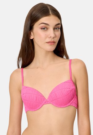 Joven con cabello largo y castaño, vistiendo un sujetador texturizado de color rosa brillante con tirantes delgados sobre un fondo liso.